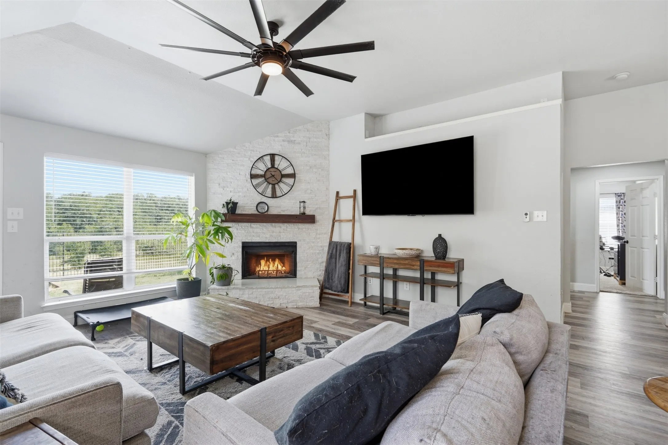 Vaulted ceiling living area featuring a stone-clad fireplace with a wood mantel, large windows with horizontal blinds, and wood-finish flooring
