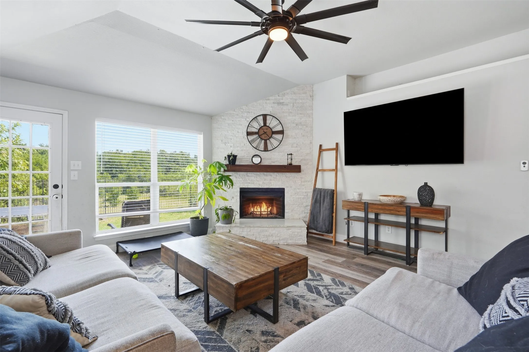 Living room featuring a vaulted ceiling with a ceiling fan, a fireplace with a stone surround, a wood mantel, and large​​‌​​​​‌​​‌‌​‌‌​​​‌‌​‌​‌​‌​​​‌​​ windows