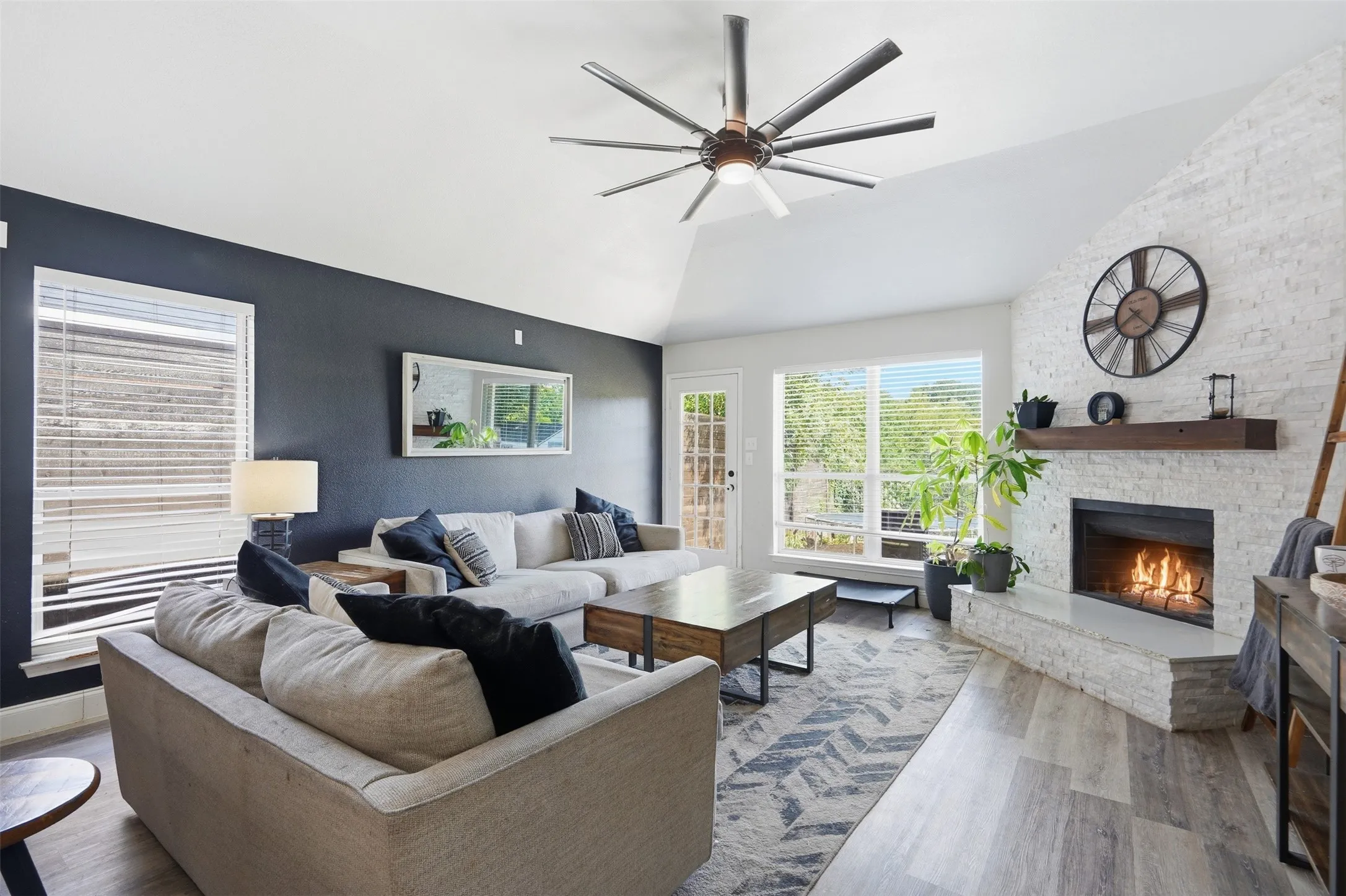 Spacious living area featuring a vaulted ceiling with a modern ceiling fan, a white brick fireplace with a wood-finish mantel, large windows with horizontal blinds, and light wood-finish flooring