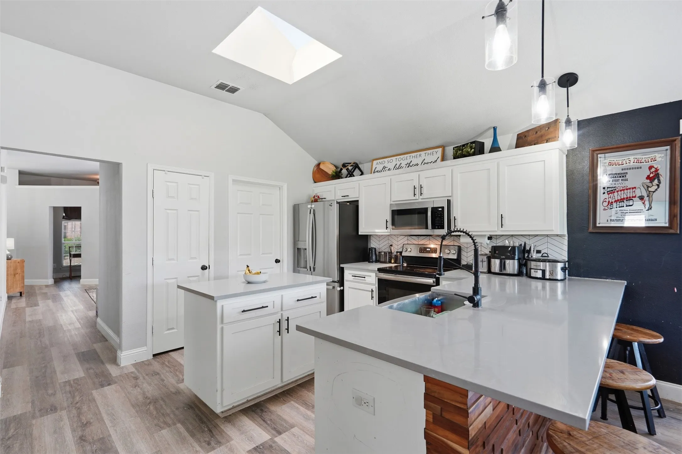 Vaulted ceiling kitchen featuring a skylight, wood-finish flooring, white cabinetry with dark hardware, stainless steel appliances, and a central island
