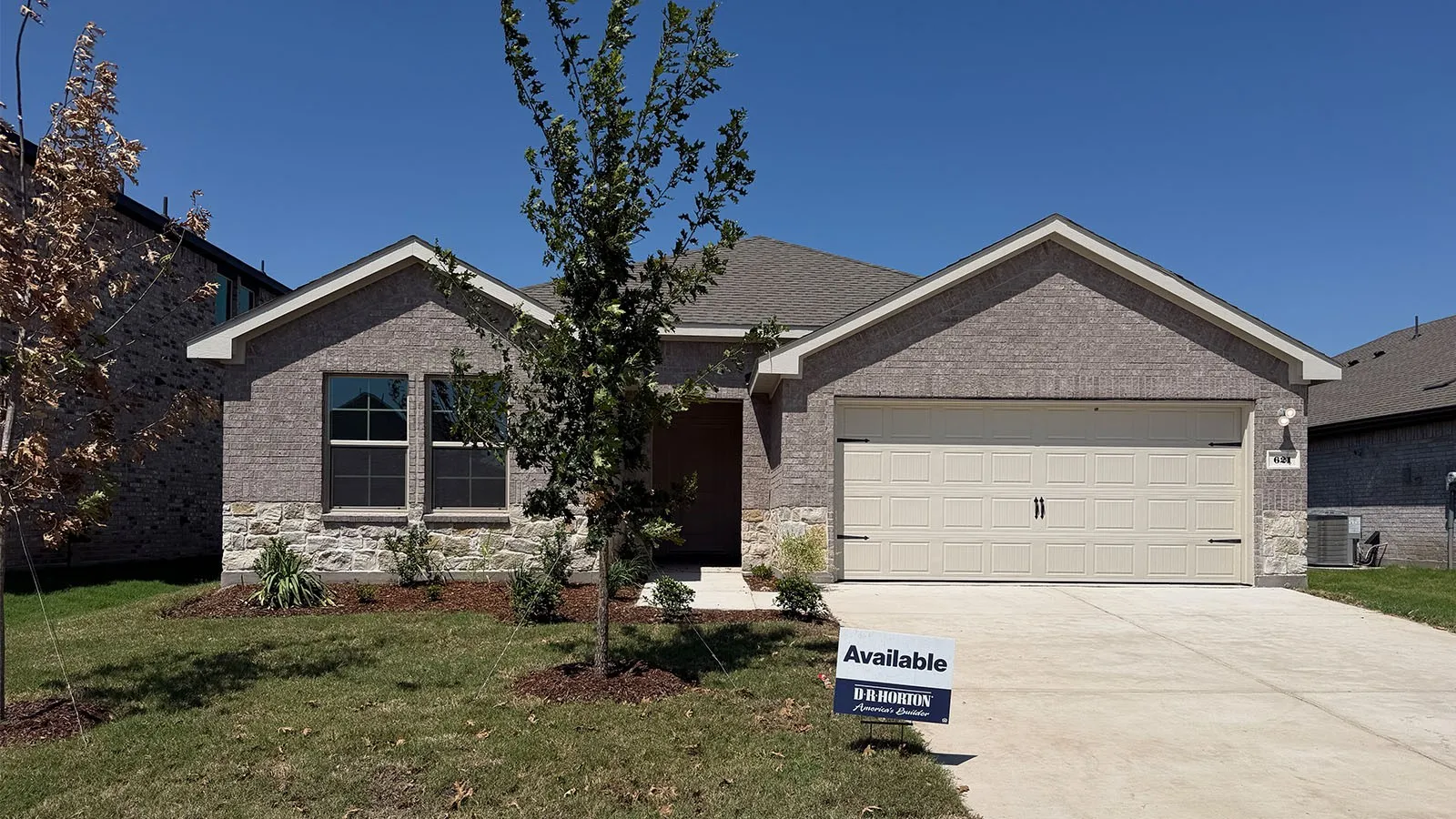 Brick single-story home featuring a two-car attached garage with panel doors, front-facing gable roof with architectural trim, and a manicured front lawn with newly planted landscaping