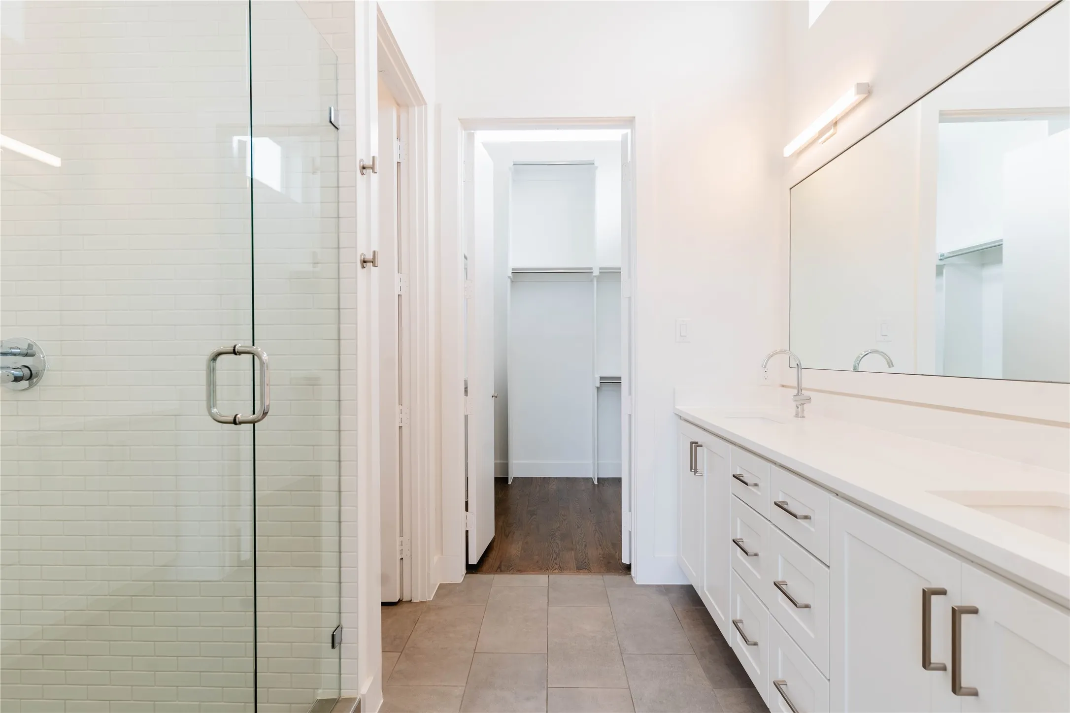 Bathroom featuring a dual-sink vanity with white cabinetry and a light-toned countertop, a frameless glass shower enclosure with subway tile surround, and neutral square tile flooring