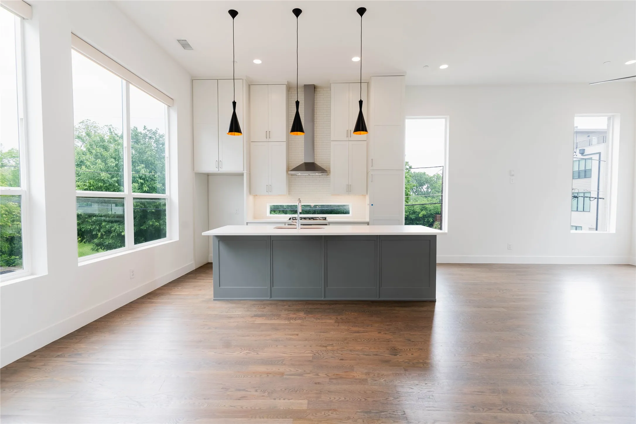 Spacious kitchen and living area featuring wood-finish flooring, a large island with a light-toned countertop, white cabinetry, and abundant natural light from multiple windows