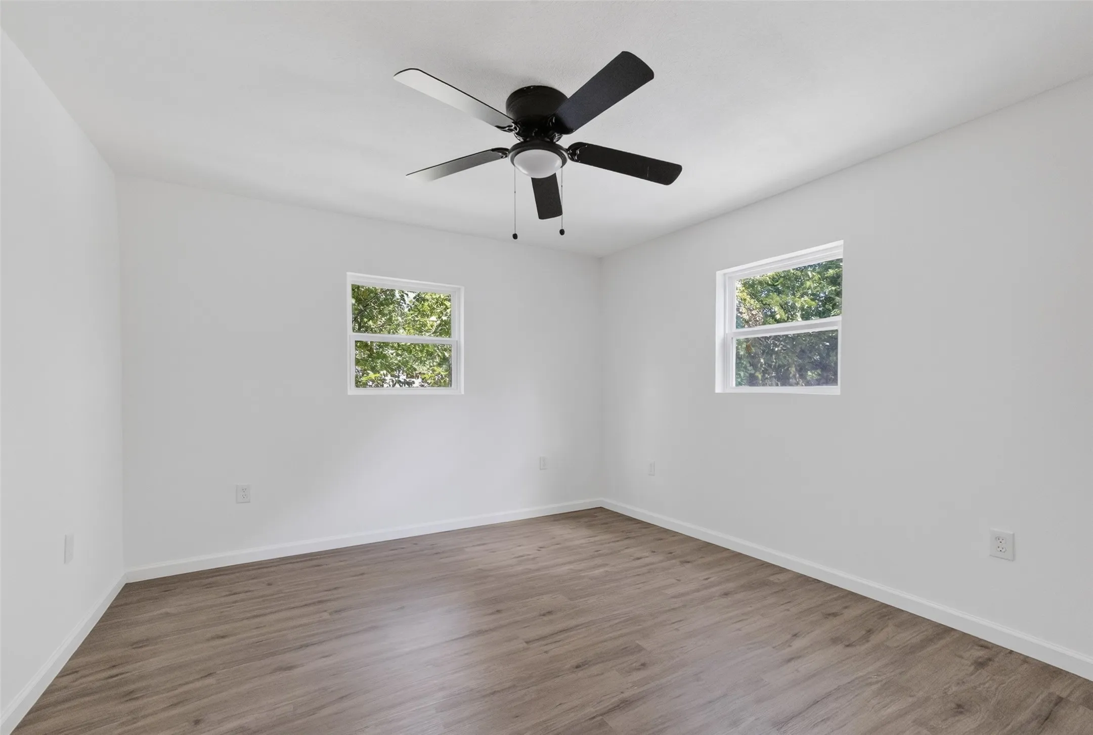 Crisp white walls frame wood-finish flooring in this room, featuring two windows and a ceiling fan with integrated lighting