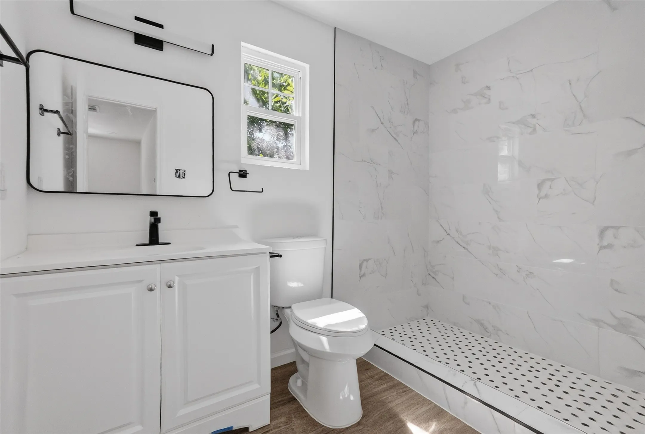 Bathroom featuring a vanity with white cabinetry and countertop, rectangular mirror with a black frame, black-finish faucet and lighting fixture, and a walk-in shower with marble-look tile walls and a mosaic tile floor