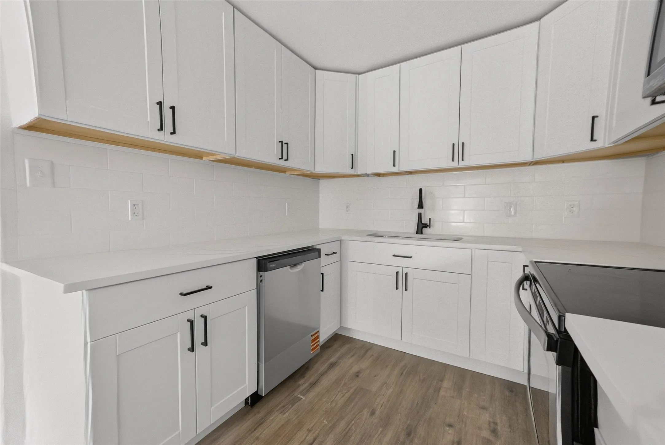 Contemporary kitchen featuring white shaker cabinetry with black hardware, white subway tile backsplash, white countertops, and wood-finish flooring