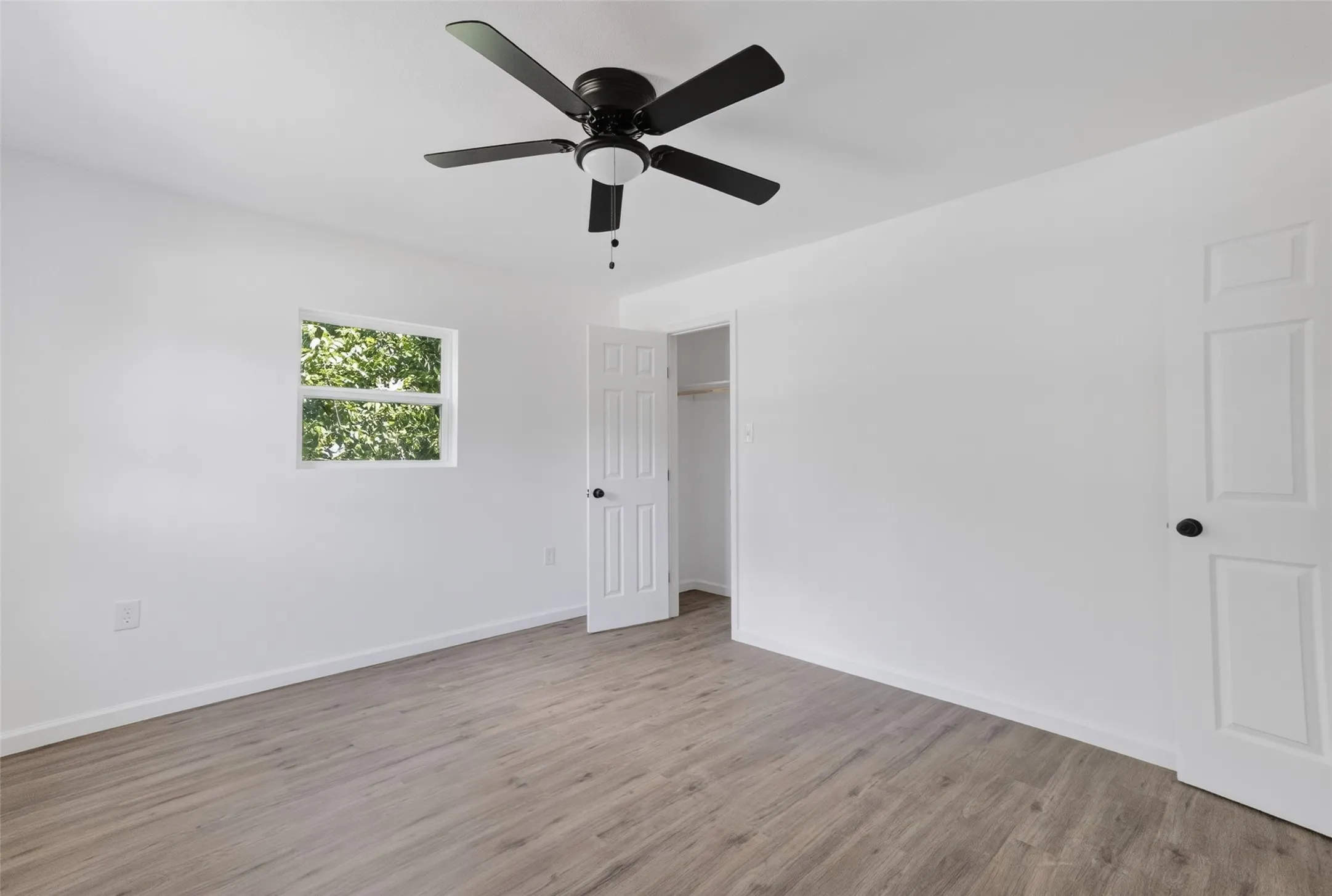 Bright room featuring wood-finish flooring, a ceiling fan with integrated lighting, a panel door with matte black hardware, and an open closet