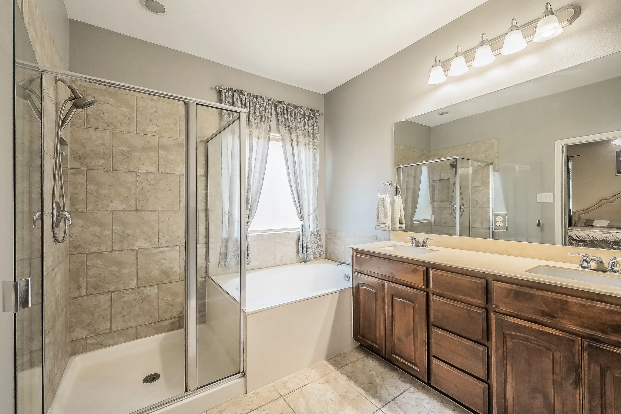 Bathroom featuring a glass-enclosed shower with tile surround, a built-in tub, a dual-sink vanity with wood-finish cabinetry, a large mirror, and tile​​‌​​​​‌​​‌‌​‌‌​​​‌‌​‌​‌​‌​​​‌​​ flooring
