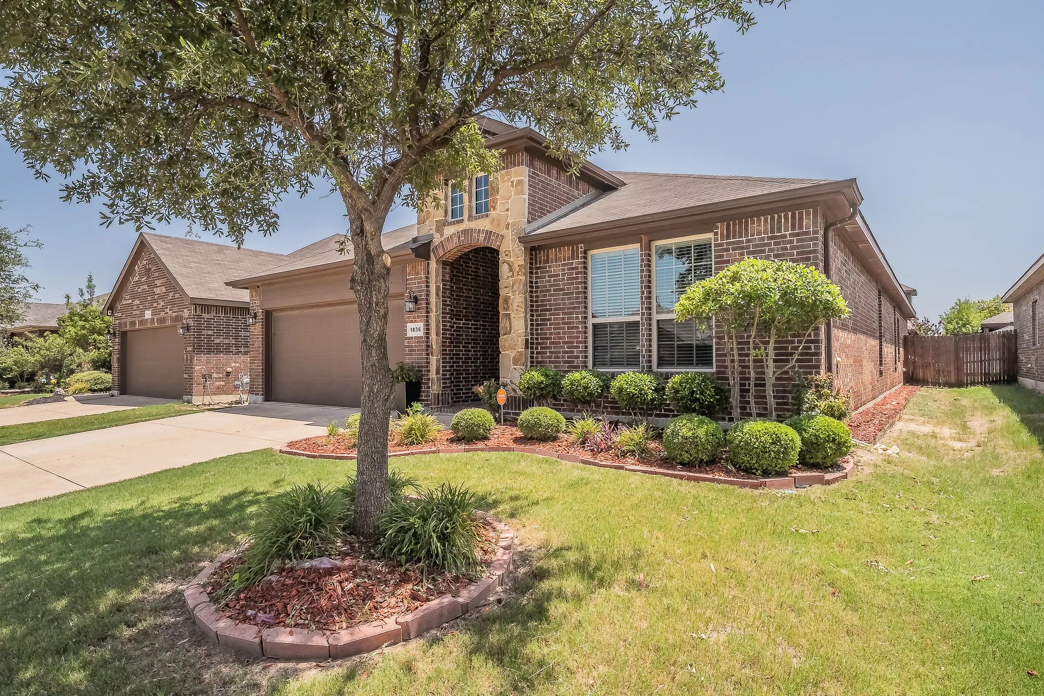 Brick and stone facade residence featuring a two-car garage, manicured shrubbery, and a mature  beautiful tree in the front yard.