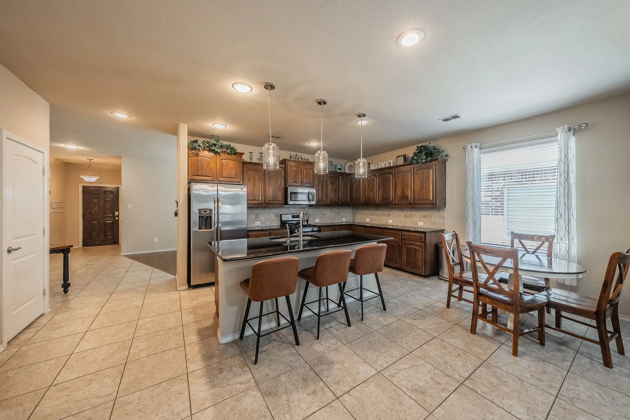 Open-concept kitchen featuring dark wood cabinetry, stainless steel appliances, a granite countertop island with pendant lighting, and tile flooring
