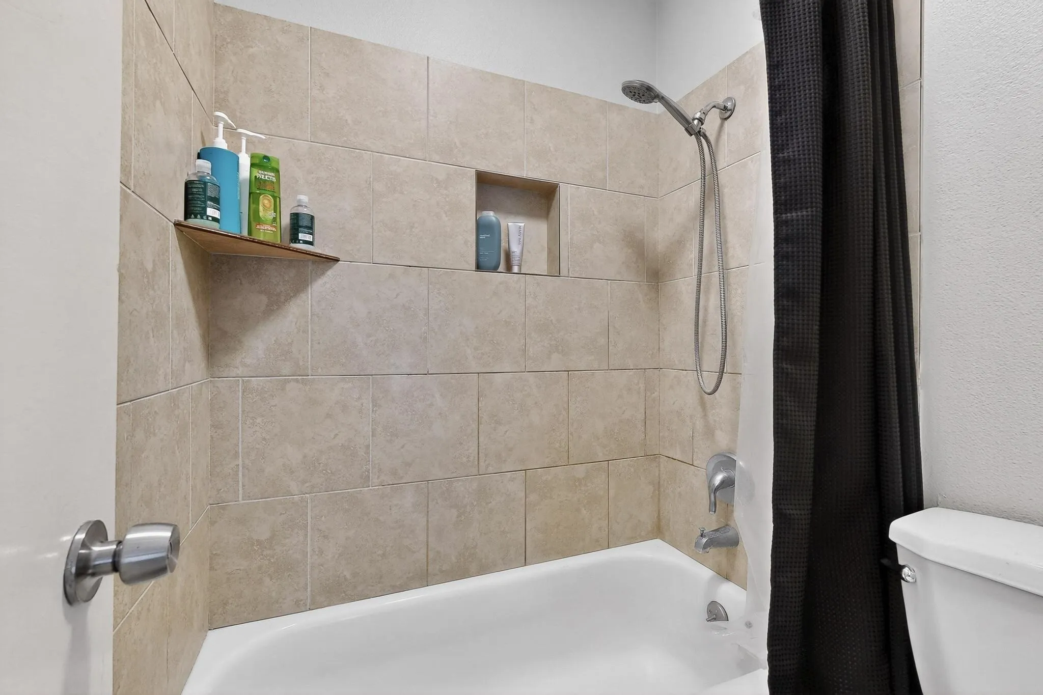Tub and shower combination featuring neutral-toned rectangular wall tiles, built-in shower niche, and an additional corner shelf
