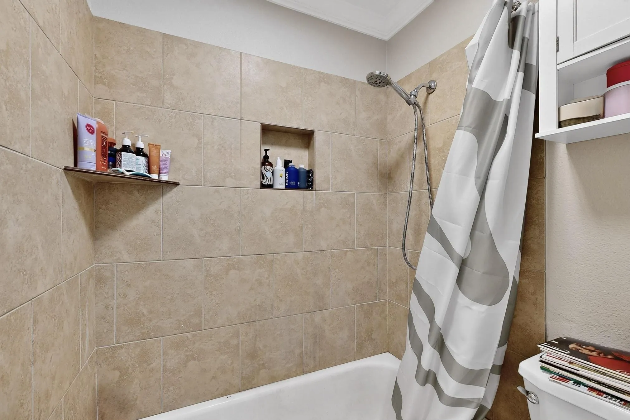 Tub shower with beige tile surround, built-in shower niche, and wall-mounted shelving