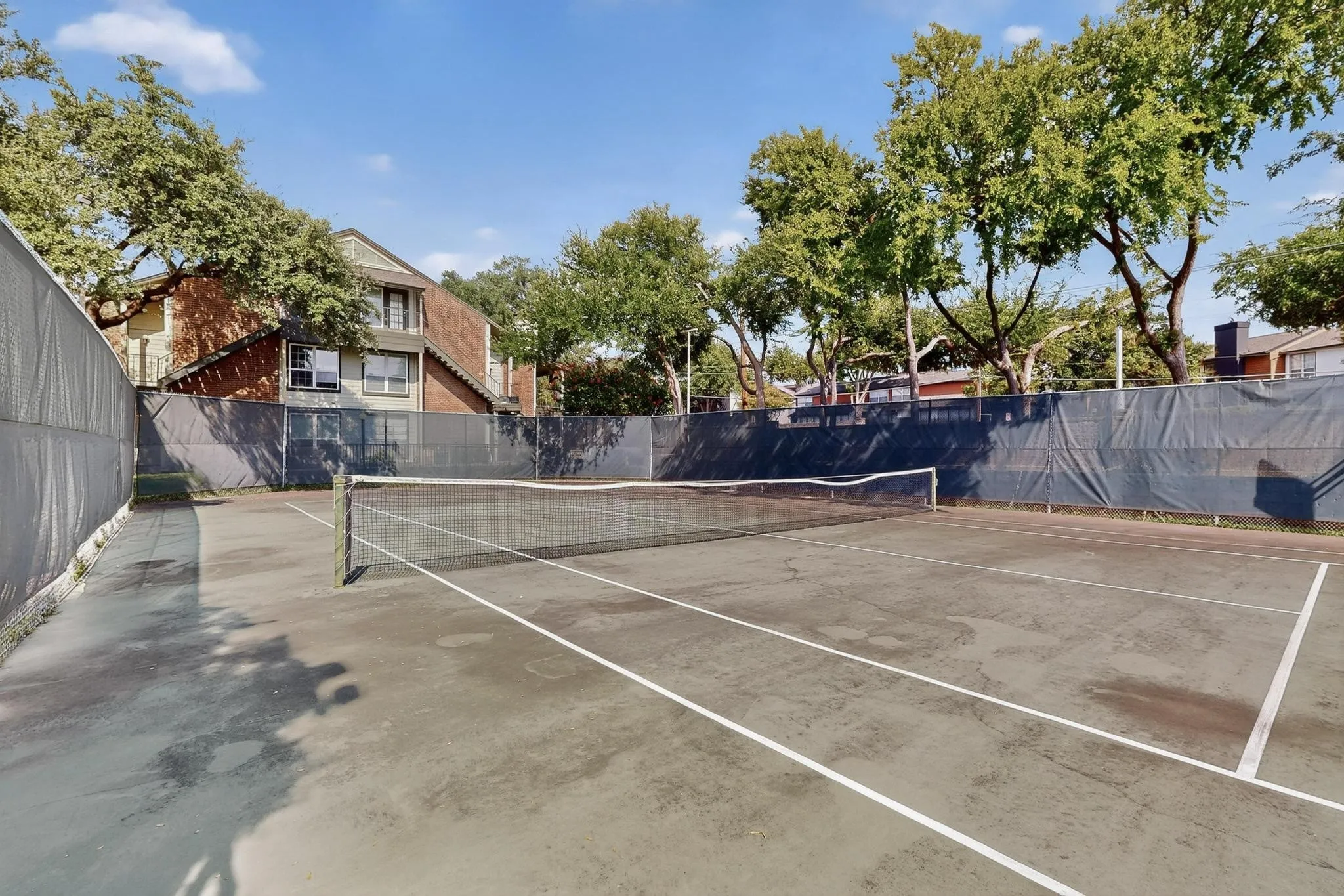 Private tennis court featuring a net and court lines, surrounded by a dark privacy screen and mature trees
