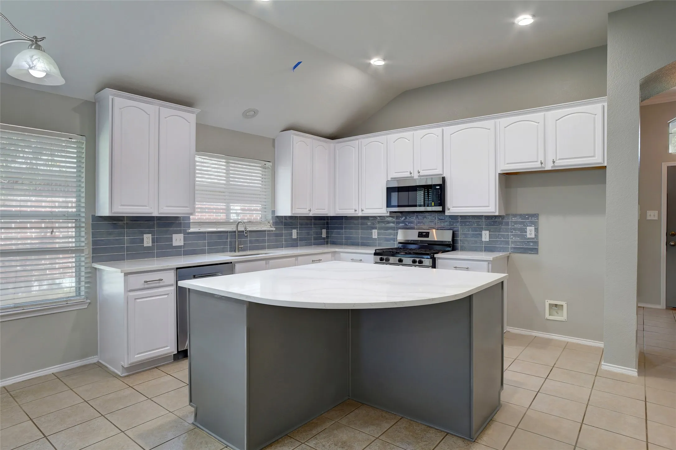 Kitchen featuring a curved central island with light-toned countertops, white cabinetry, a blue tile backsplash, stainless steel appliances, and tile flooring