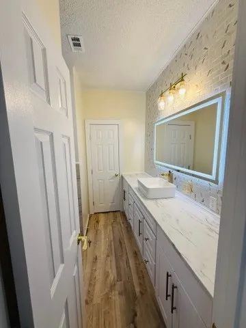 Bathroom with wood-finish flooring, a long white vanity with multiple cabinets and drawers, a vessel sink, a white countertop, and a vanity mirror with integrated lighting