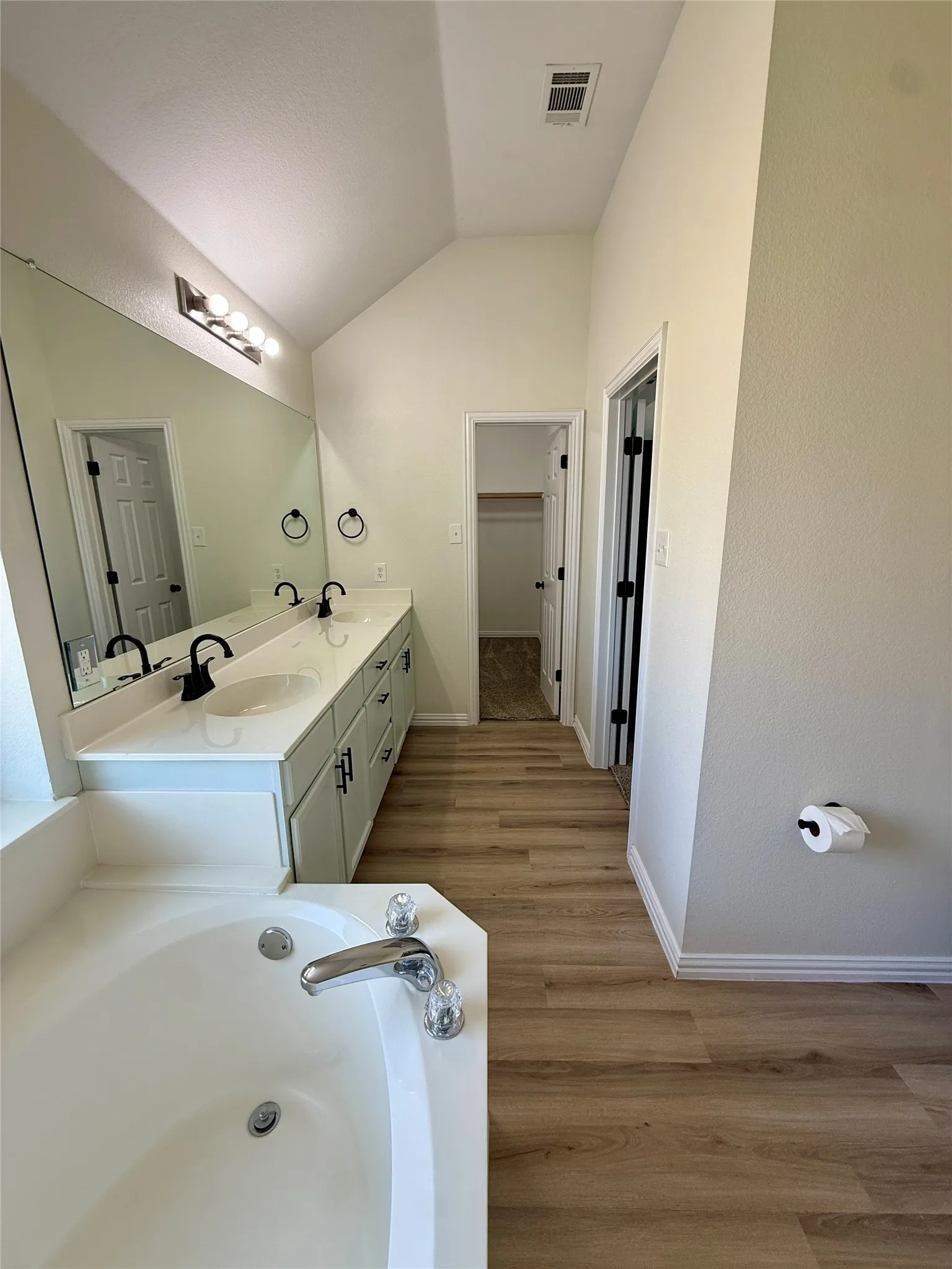 Bathroom featuring a built-in soaking tub, dual vanity with white countertop, matte black fixtures, wood-finish flooring, and vaulted ceiling