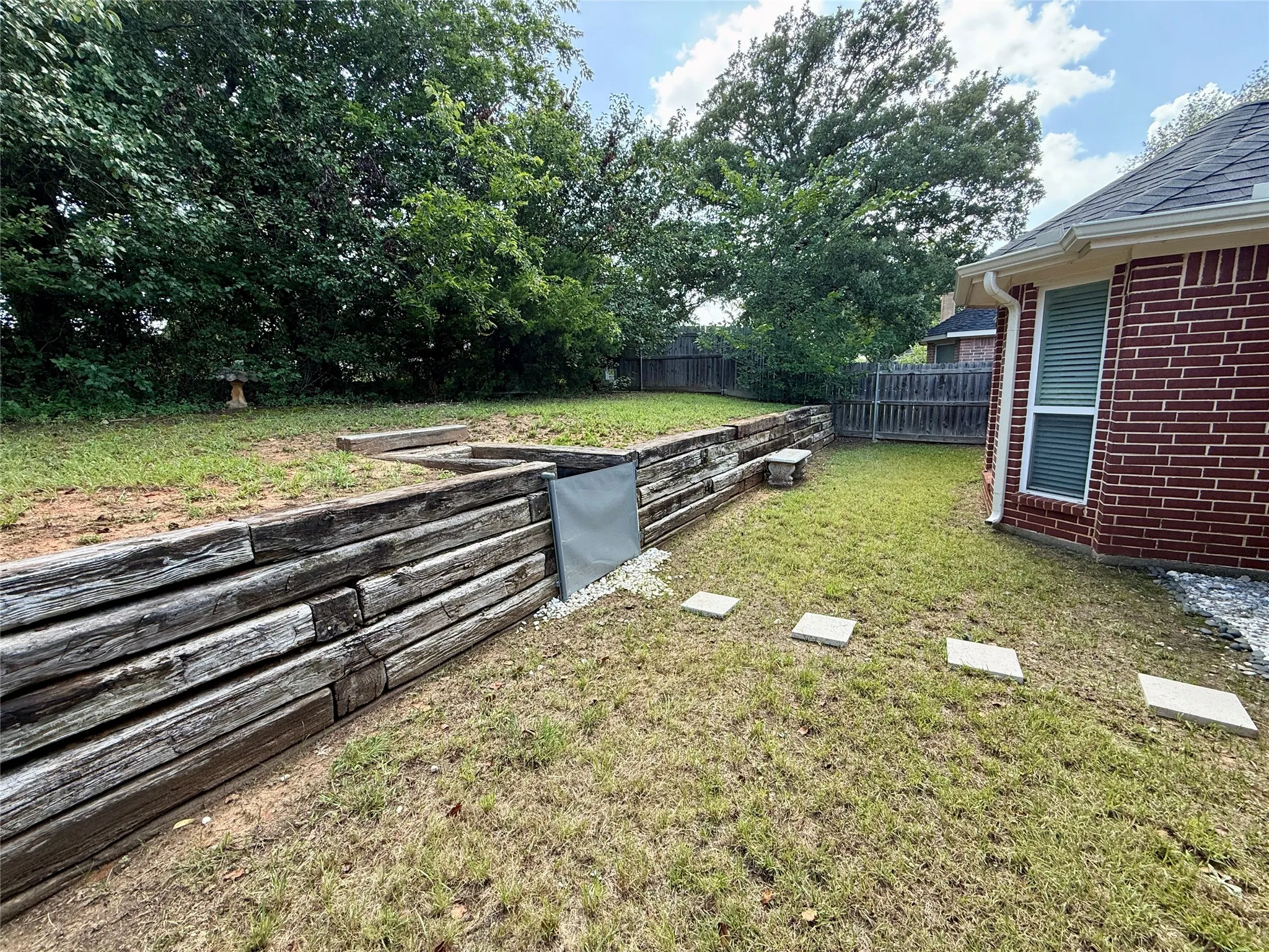 Tiered backyard featuring a retaining wall constructed from stacked timber, a manicured lawn, and mature perimeter trees