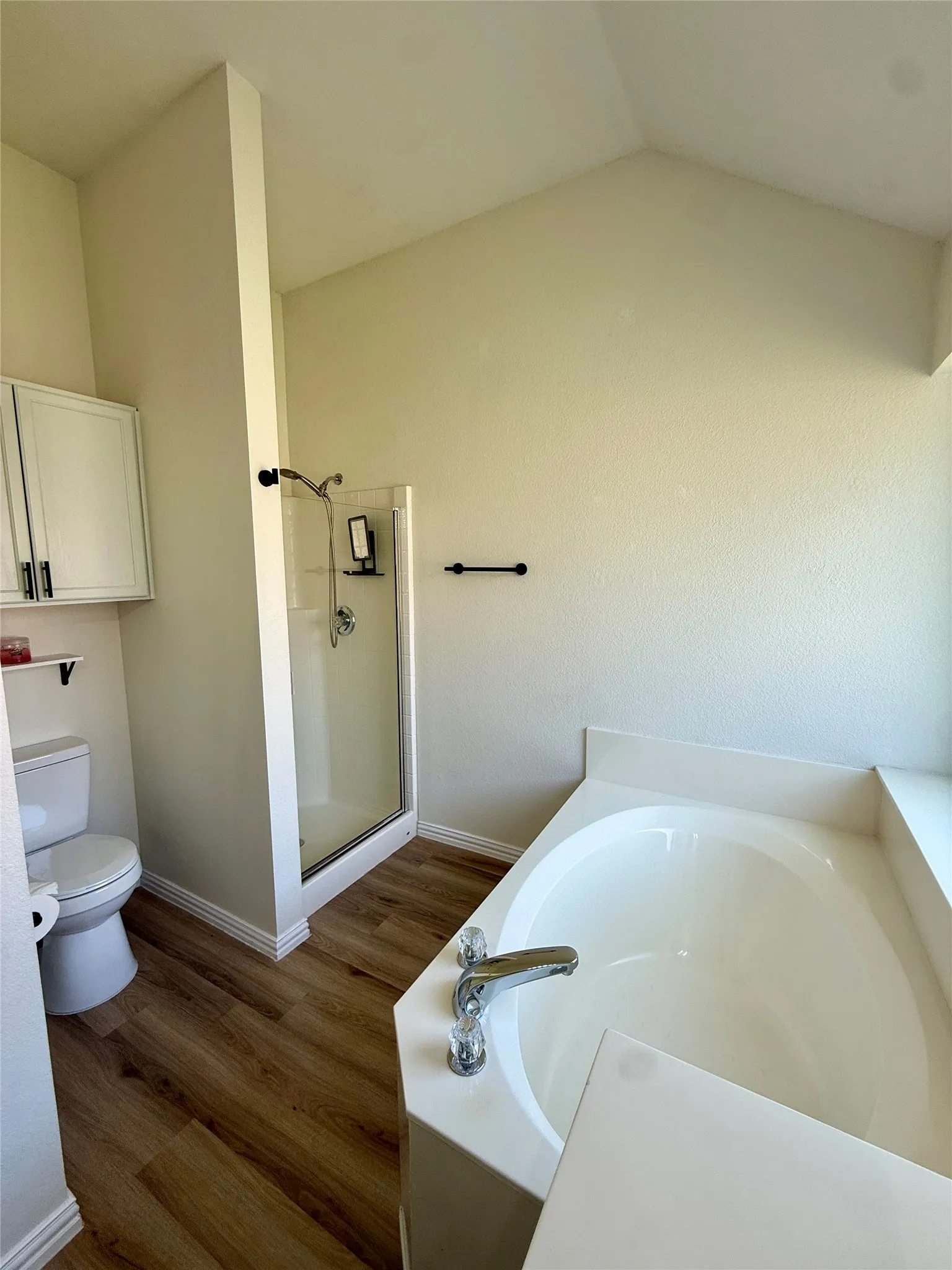 Corner soaking tub with chrome fixtures, glass-enclosed shower, wood-finish flooring, white cabinetry, and vaulted ceiling