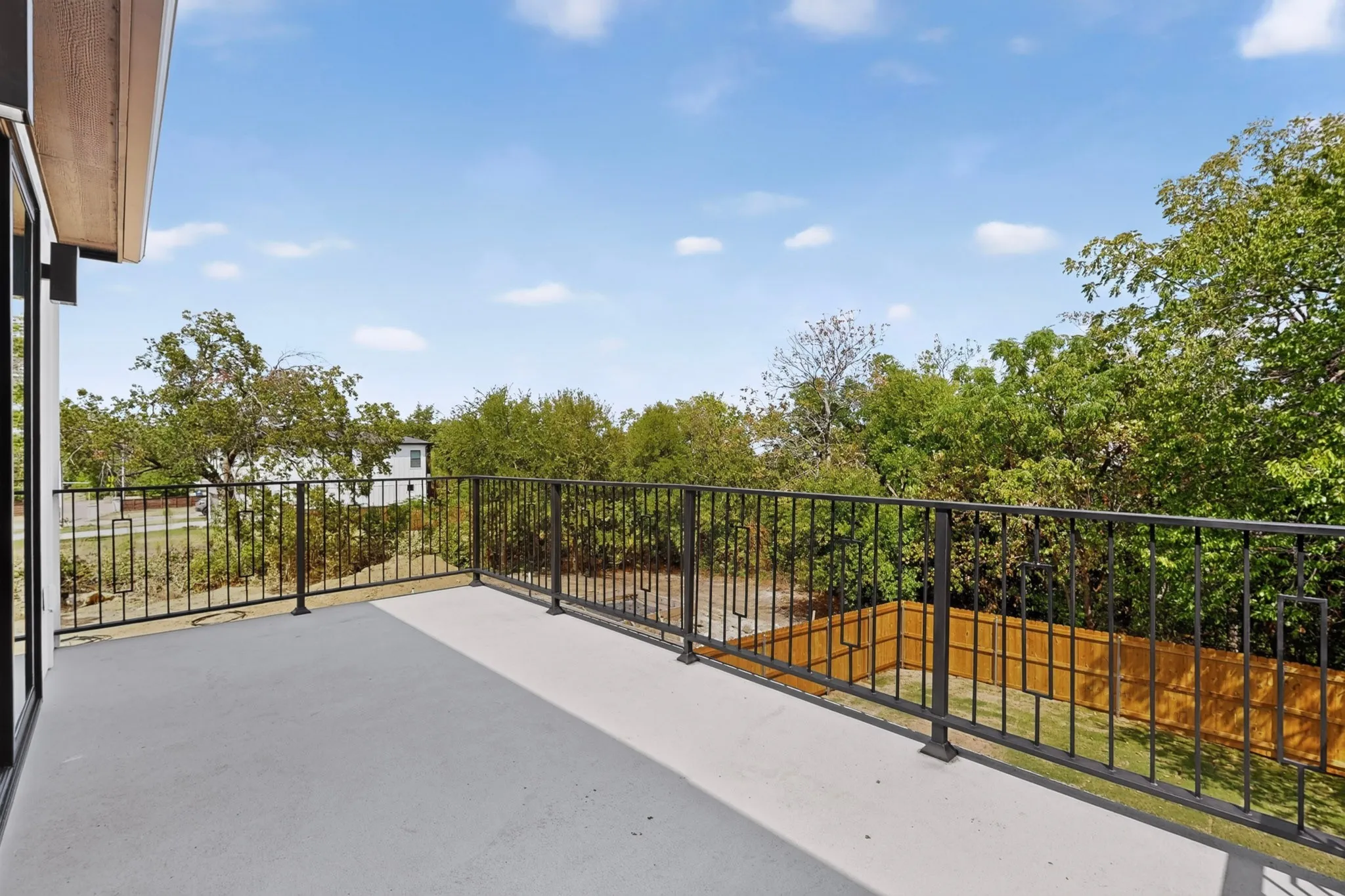 Expansive outdoor balcony with a black metal railing and concrete flooring, overlooking mature trees and a wood fence
