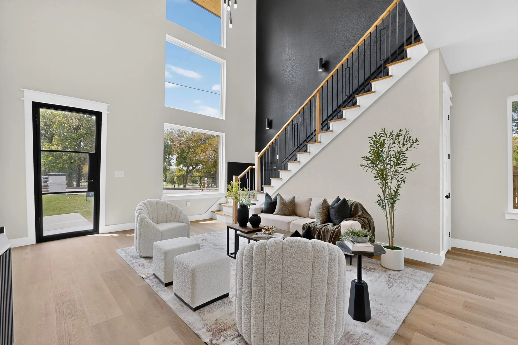 Double-height ceiling, large multi-pane windows, wood-finish flooring, staircase with metal balusters and wooden handrail, and a black-framed glass entry door