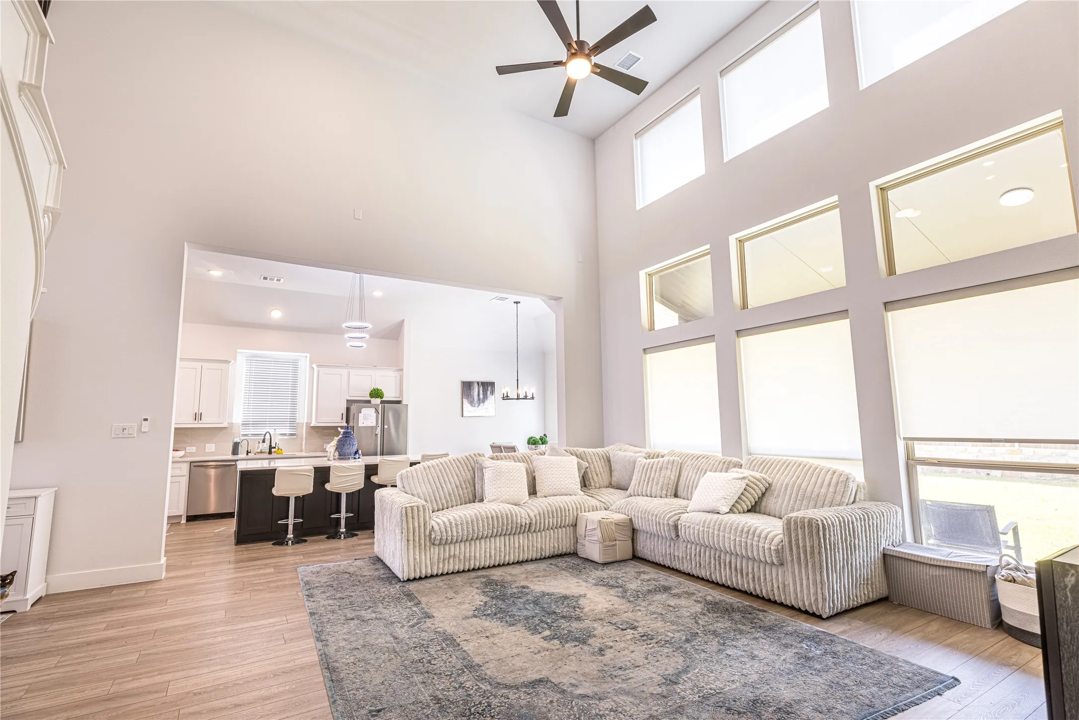 Double-height ceiling living space featuring a contemporary ceiling fan, expansive multiple-pane windows, wood-finish flooring, and an open concept layout connecting to a kitchen with an island and pendant lighting