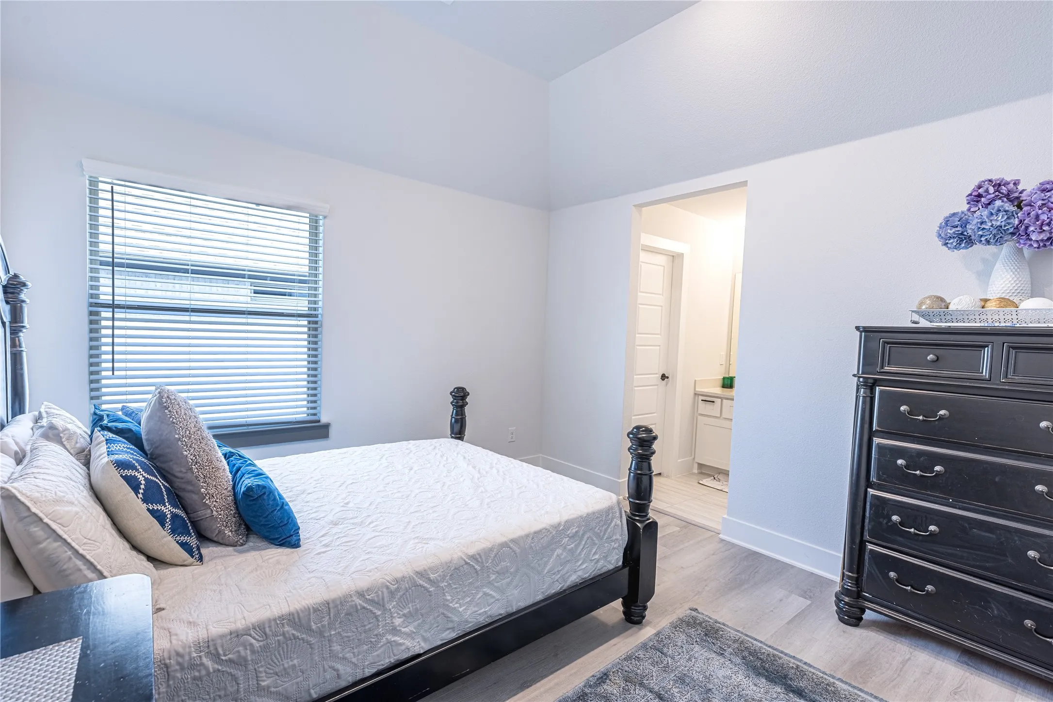 Vaulted ceiling bedroom with light wood-finish flooring, white walls, and a window featuring blinds