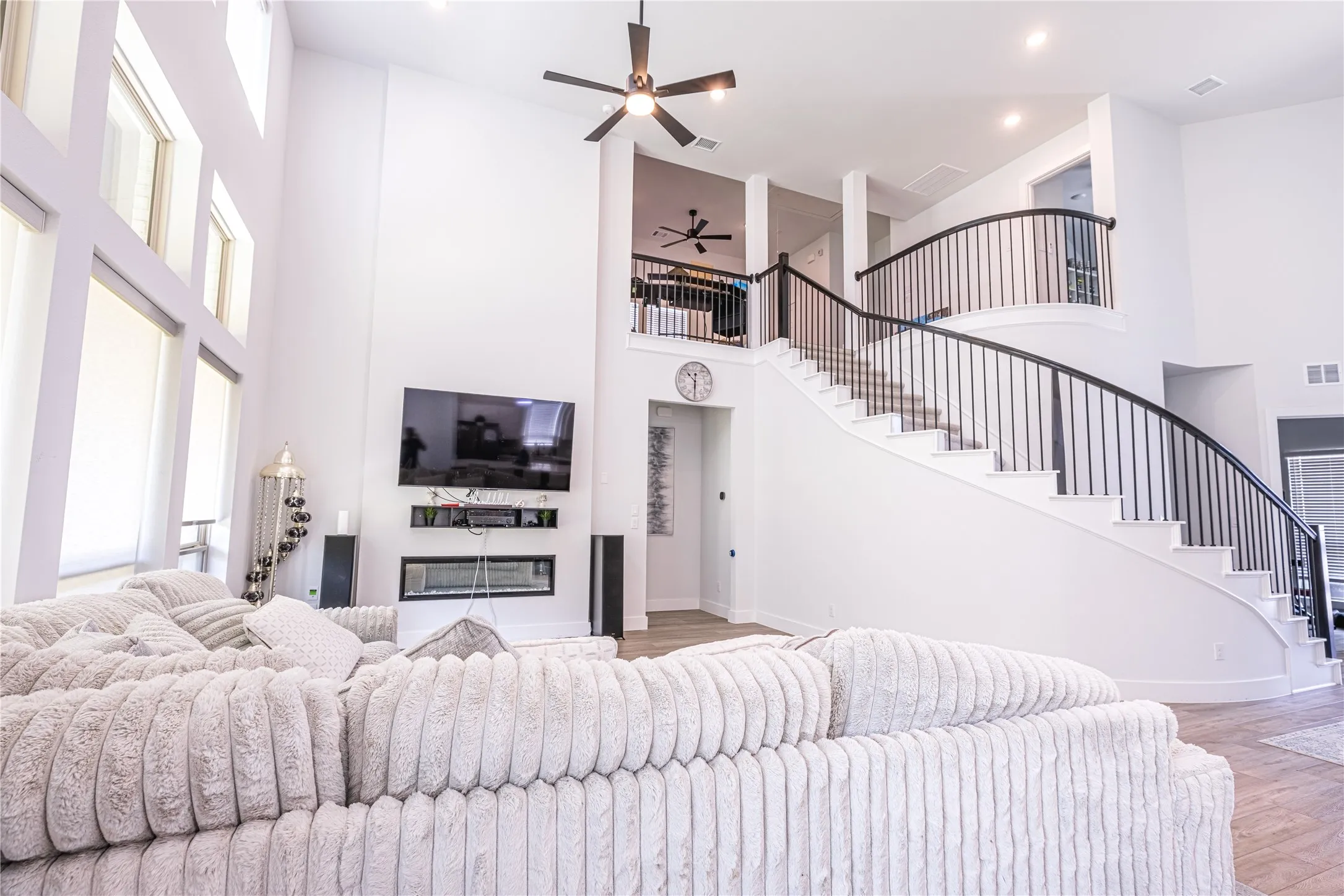 Two-story living area featuring a curved staircase with black metal balusters, high ceilings, large windows with white frames, and a built-in electric fireplace
