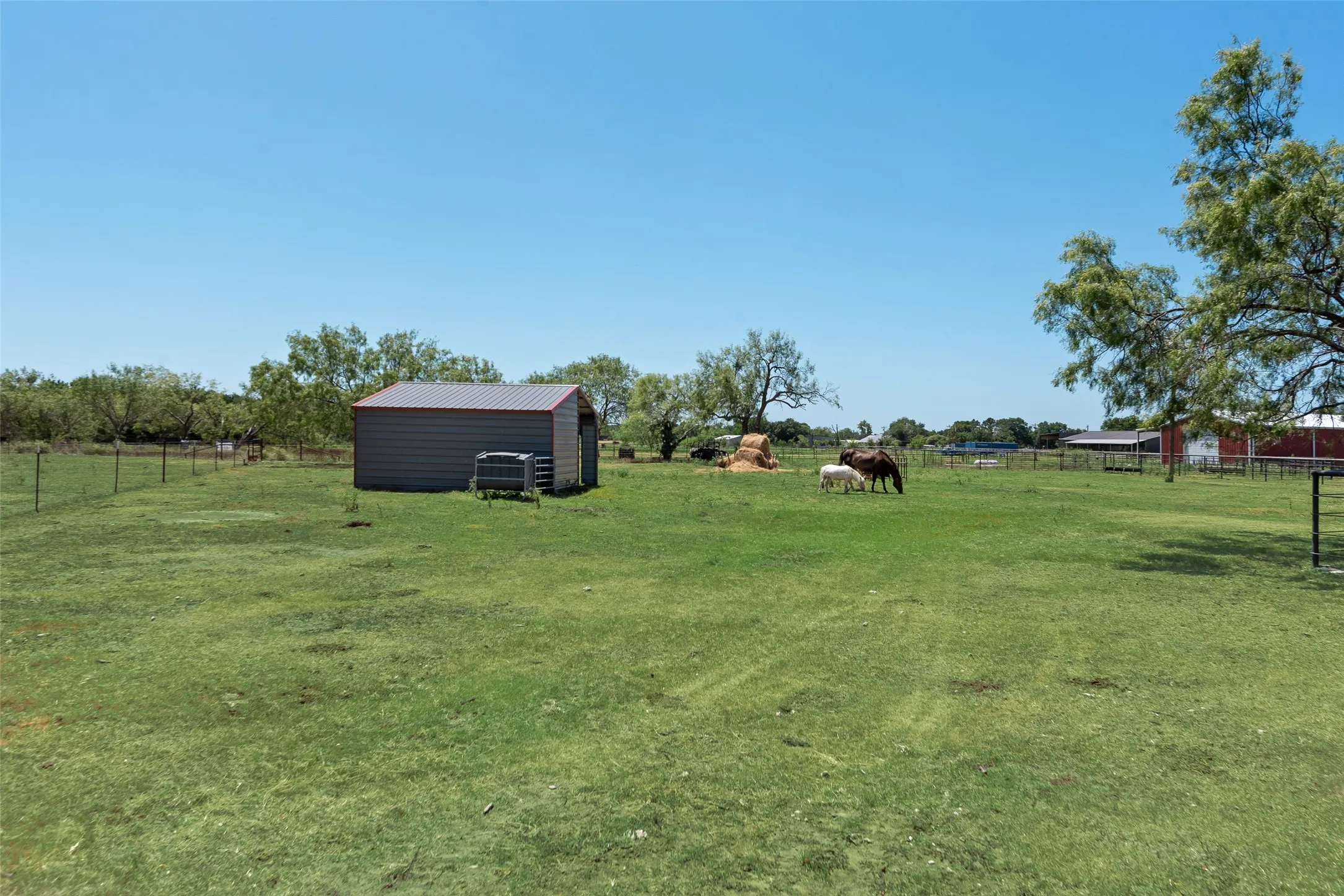 Spacious green acreage featuring a metal shed building with a red trim roof, wire perimeter fencing and scattered mature trees.