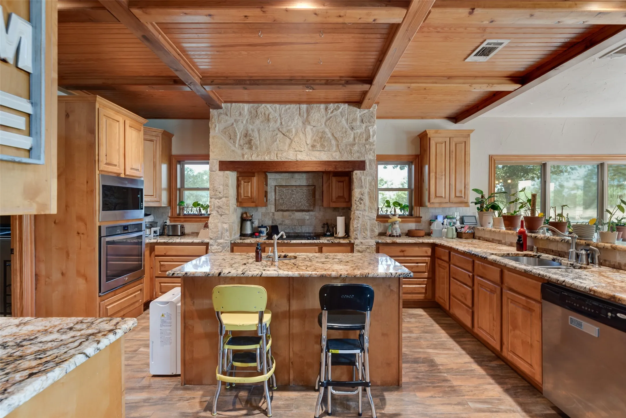 Spacious kitchen featuring a central island with a stone-finish countertop, extensive wood-finish cabinetry, stainless steel appliances, a stone-accented range hood, and a wood-paneled ceiling with exposed beams