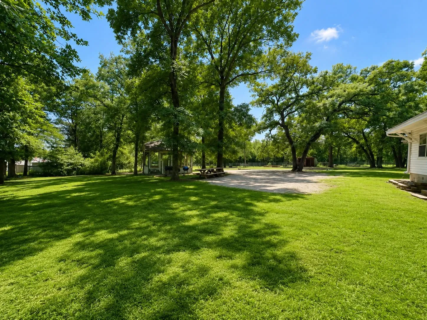 Expansive lawn with mature shade trees, a white gazebo, and a gravel parking area