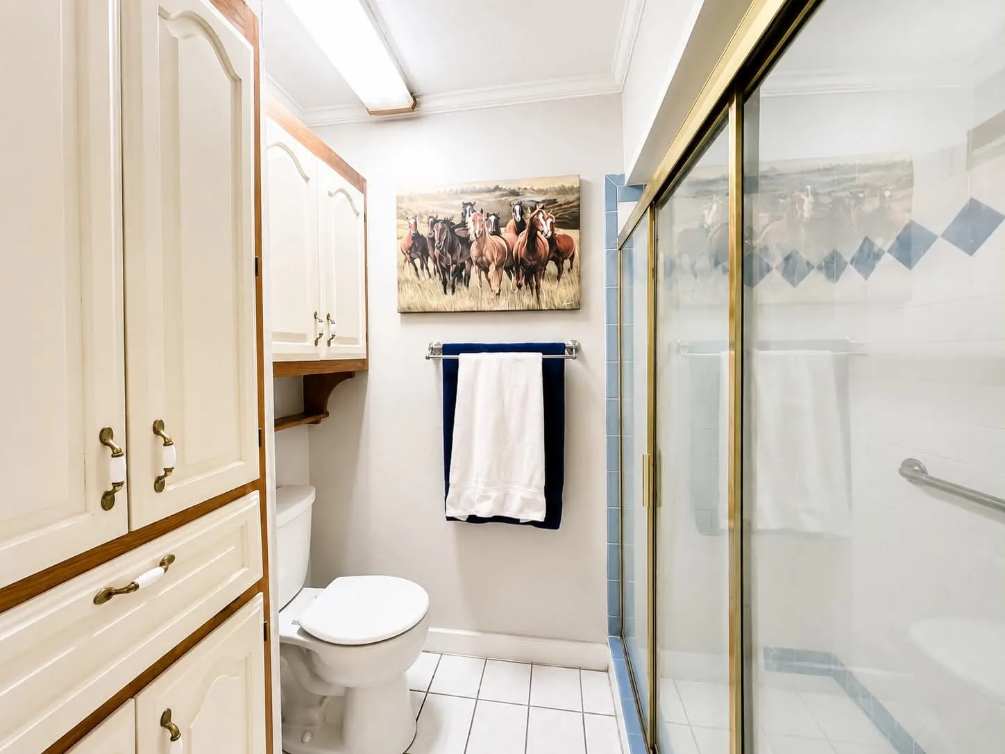 White tile flooring bathroom featuring a built-in linen cabinet with cream-colored doors and wood-finish trim, a toilet, a gold-framed glass shower enclosure, and white tiled shower walls with blue diamond accents
