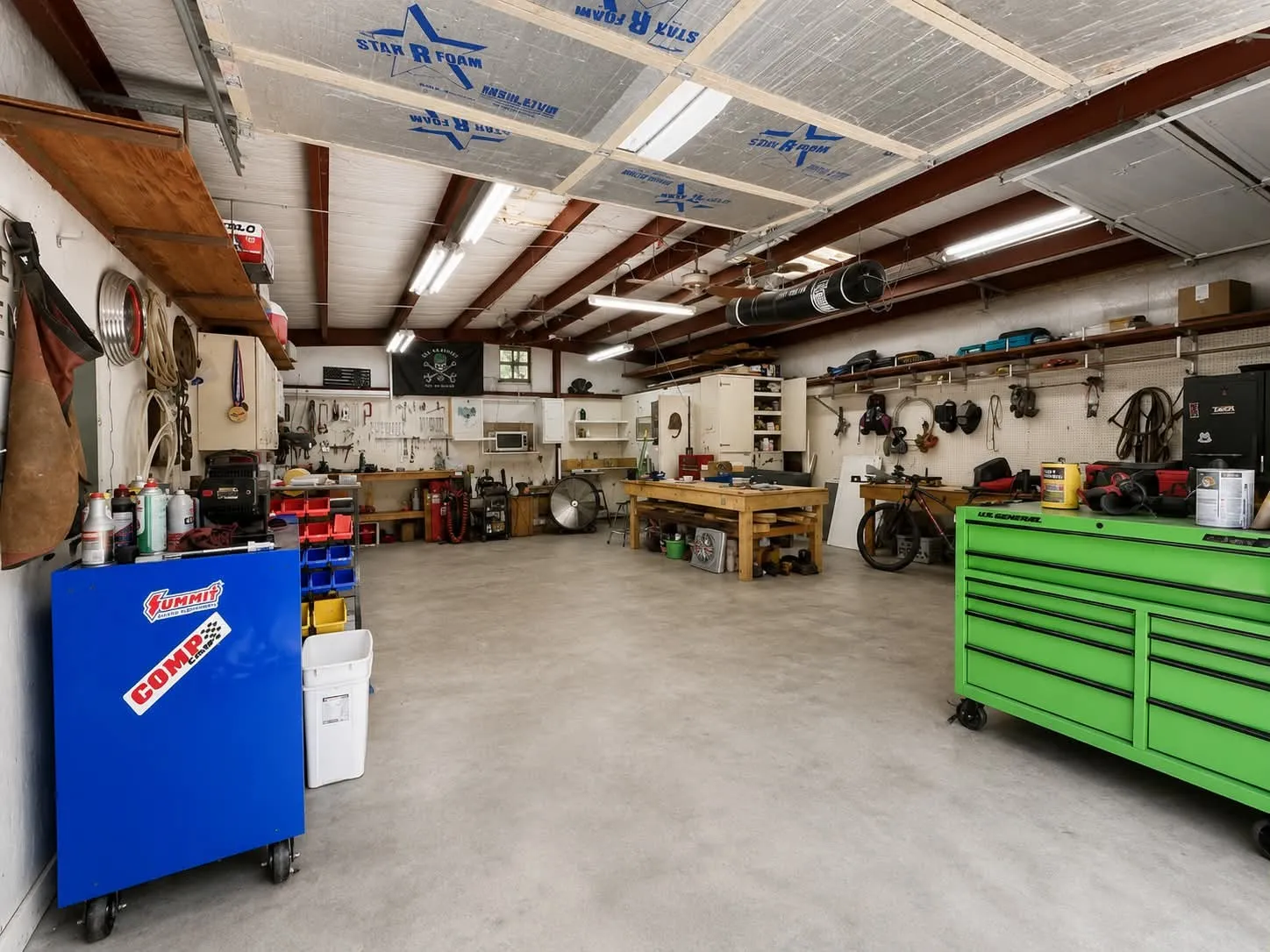 Spacious workshop area featuring a concrete floor, exposed beam ceiling with insulation panels, fluorescent lighting, extensive wall shelving, and a built-in workbench