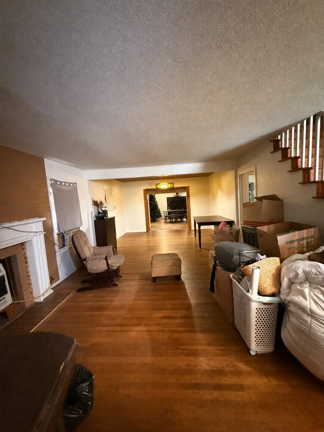 Spacious living area featuring wood-finish flooring, a traditional brick fireplace with a white mantel, and a staircase with open risers and wood handrails