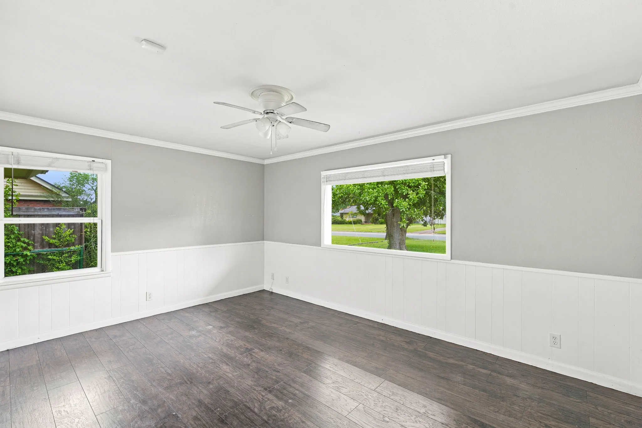 Room featuring wood-finish flooring, two windows, two-tone walls with white wainscoting, and a ceiling fan with integrated lighting
