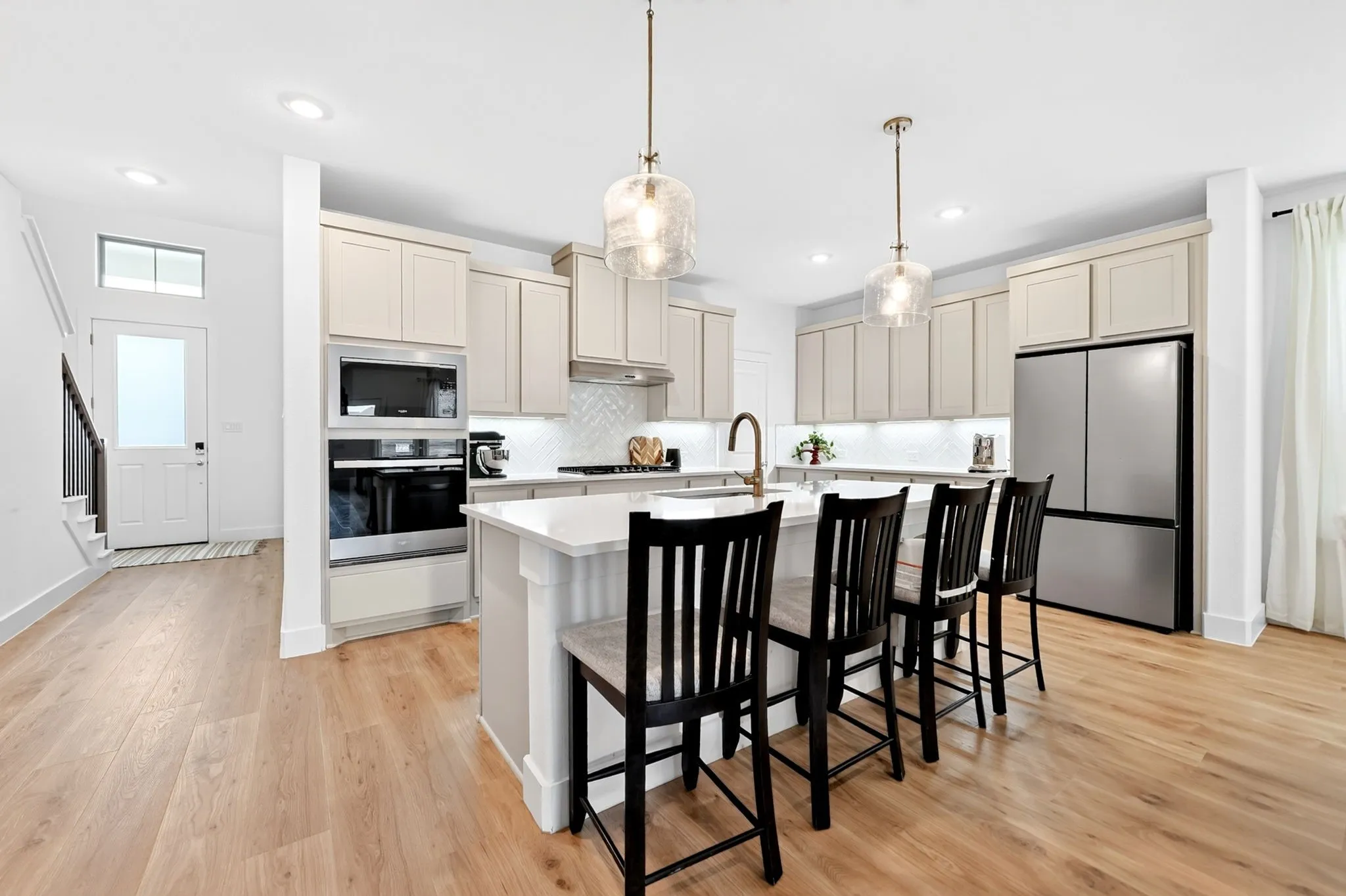 Contemporary kitchen featuring a large central island with seating, light wood-finish flooring, built-in stainless steel appliances, white shaker-style cabinetry, and pendant lighting