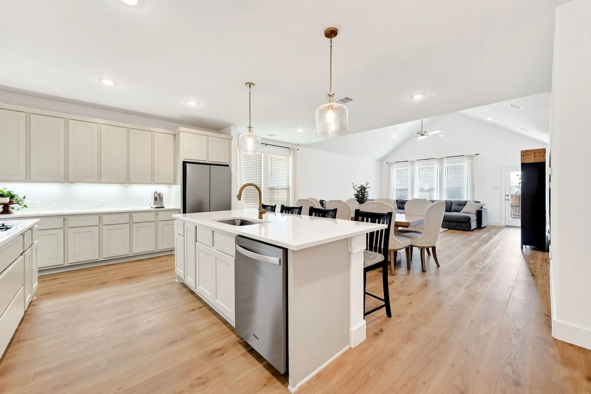 Open-concept kitchen featuring an island with a sink and pendant lighting, white cabinetry, integrated stainless steel appliances, wood-finish flooring, and a vaulted ceiling