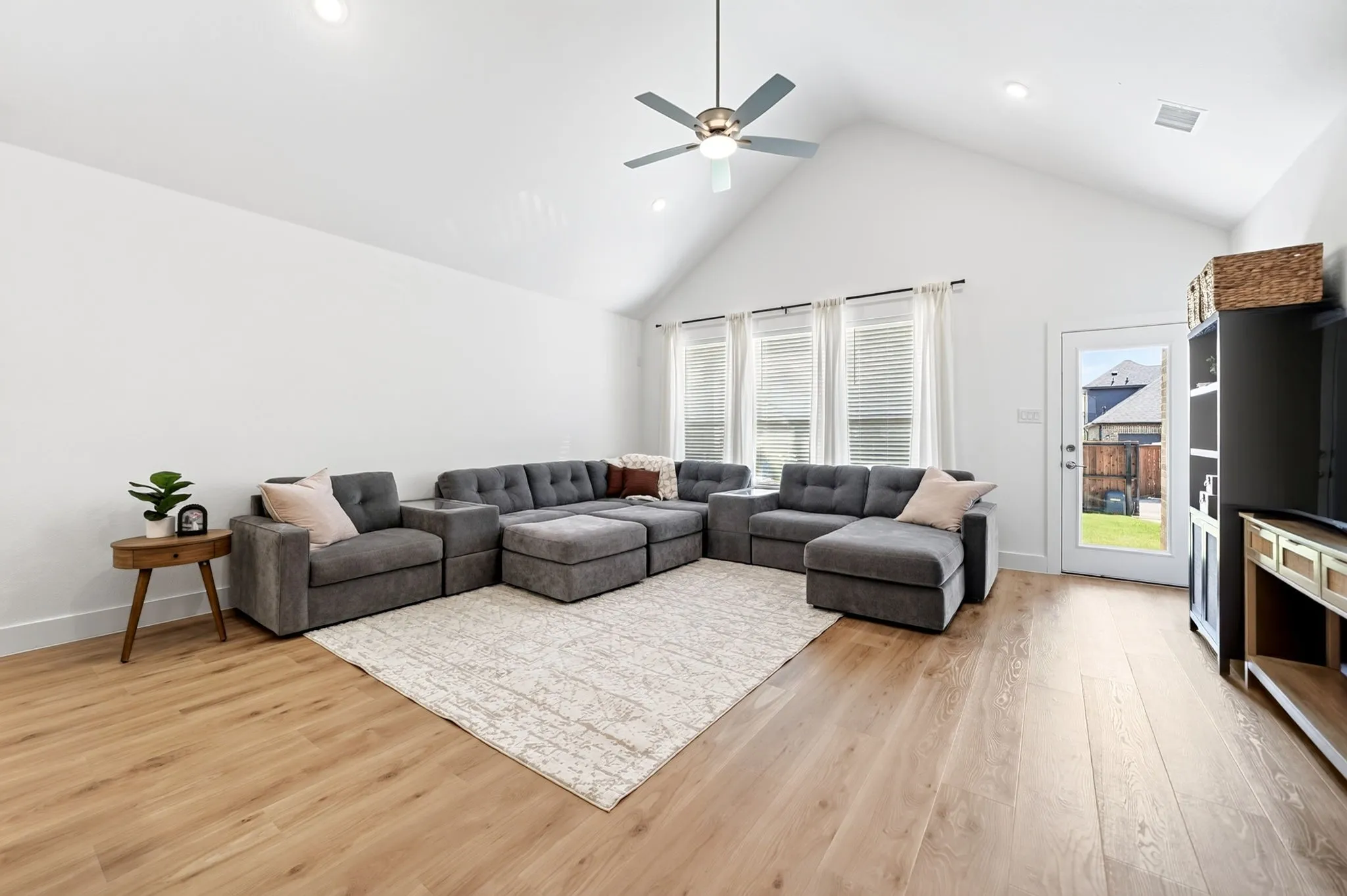 Vaulted ceiling living area featuring light wood-finish flooring, recessed lighting, a modern ceiling fan, three windows with blinds and curtains, and a glass-paneled back door