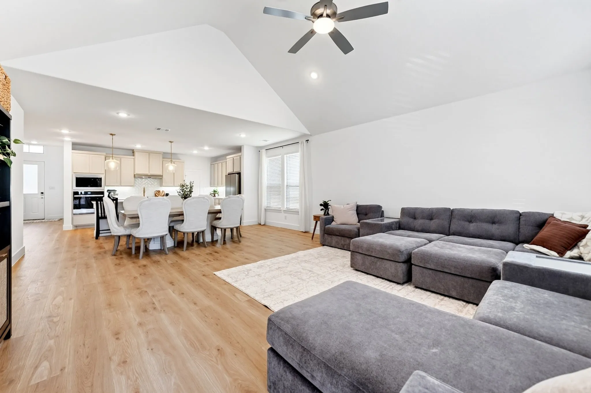 Open-concept living area featuring a vaulted ceiling with a ceiling fan, light wood-finish flooring, recessed lighting, and large windows with blinds