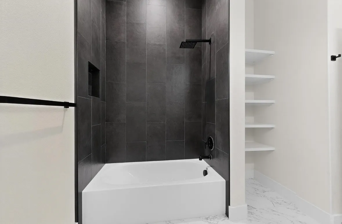 Bathroom featuring a white soaking tub with dark-toned tile surround, matte black shower fixtures, a recessed shower niche, and built-in shelving