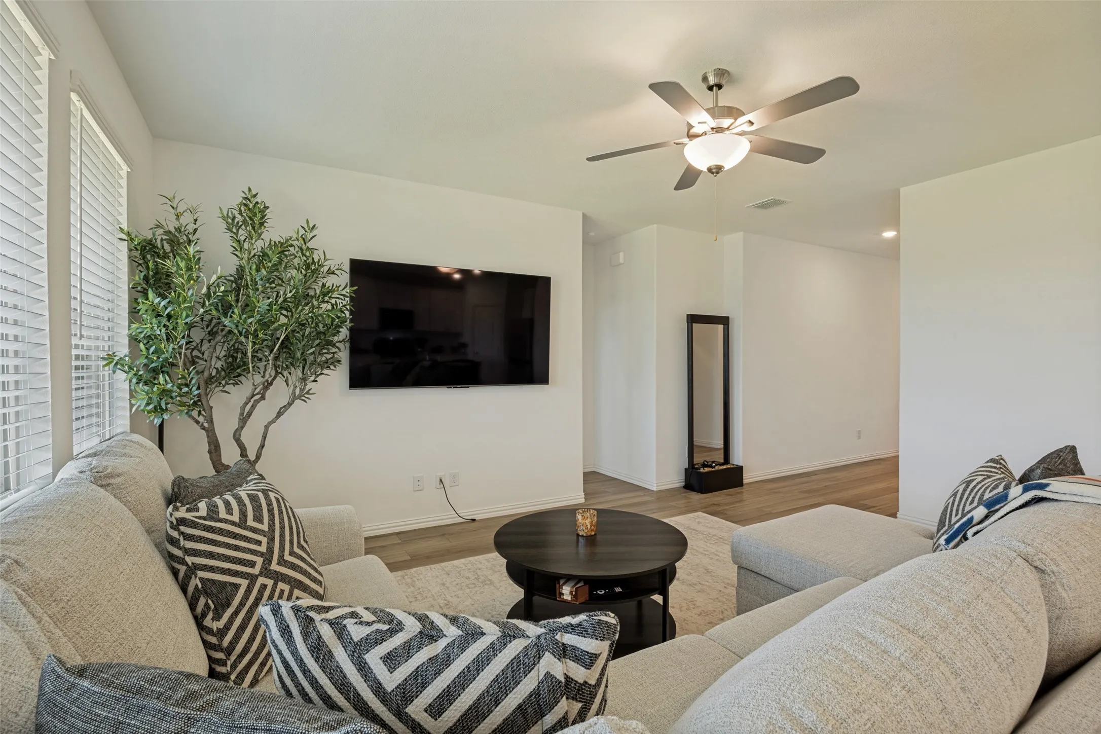 Open-concept living space featuring wood-finish flooring, white walls, a ceiling fan with integrated lighting, and multiple windows with blinds