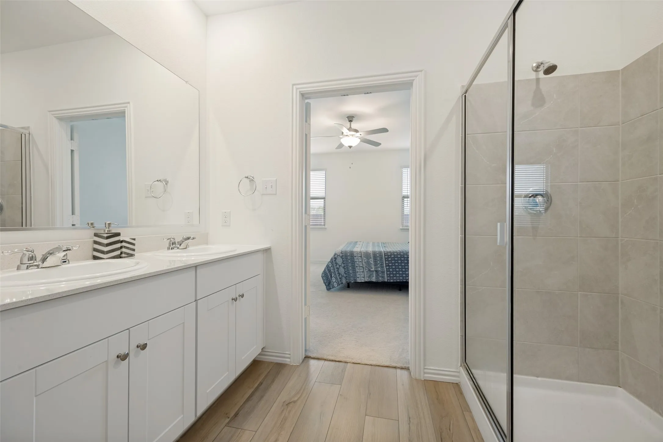 Dual vanity with white cabinetry, wood-finish flooring, a frameless glass shower enclosure, and tiled shower walls