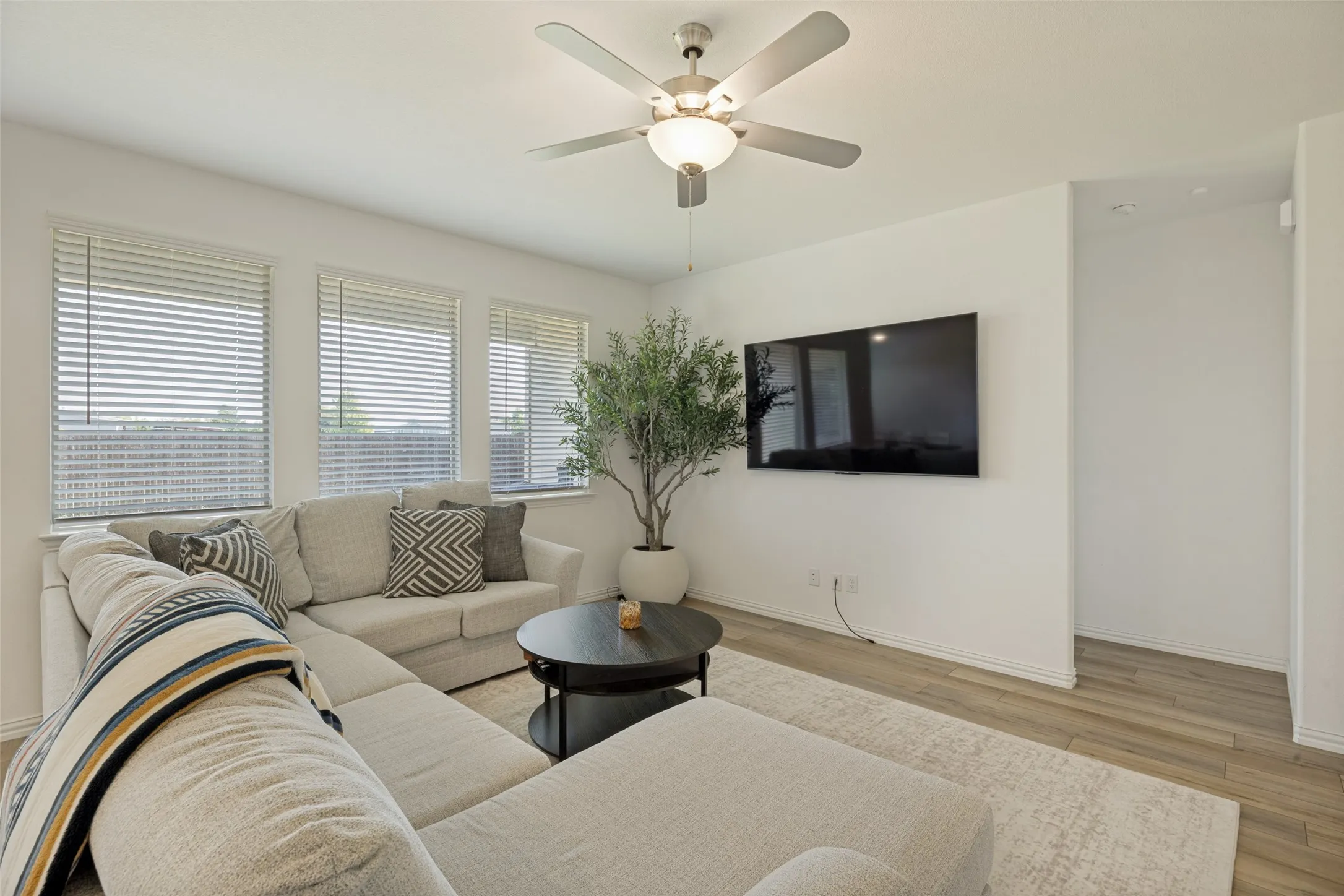Living area featuring wood-finish flooring, a ceiling fan with integrated lighting, and multiple windows with blinds