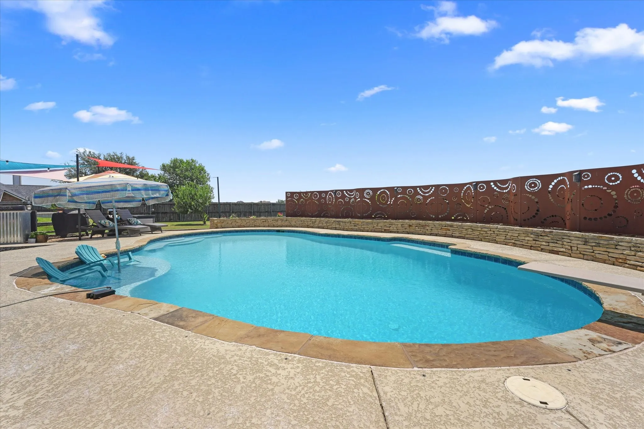 In-ground swimming pool with an art deco steel privacy wall, stone retaining wall, concrete patio, and integrated pool steps