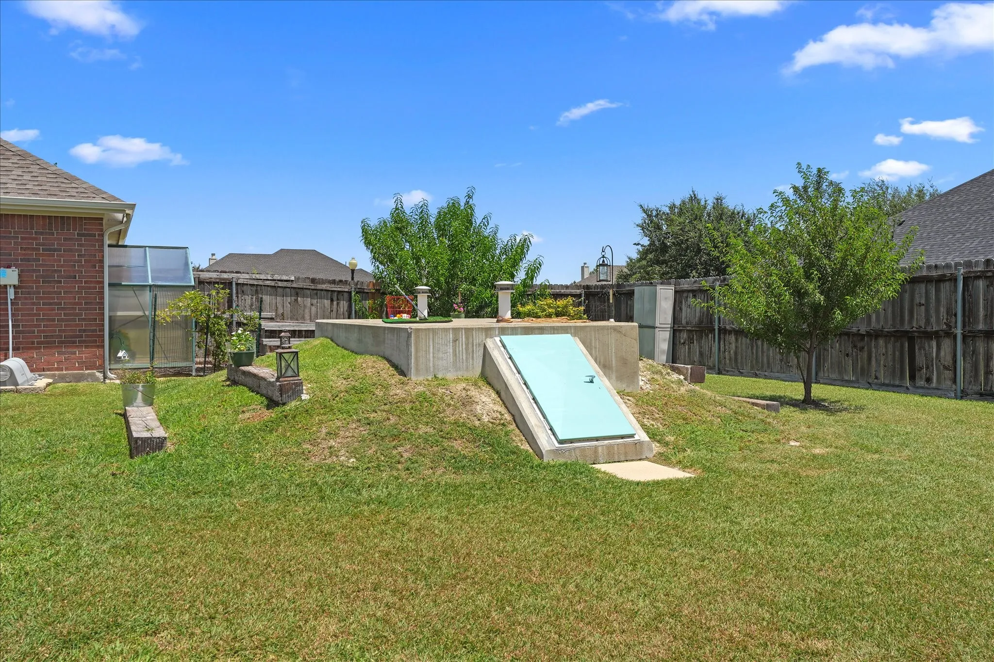 Walk-in storm shelter with a blue entrance door, a greenhouse structure, and a wood privacy fence