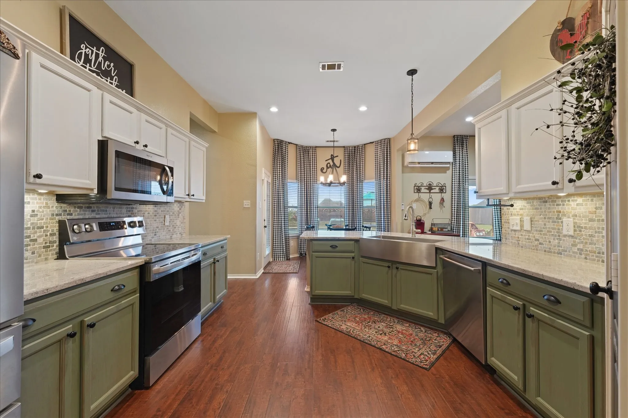 Spacious kitchen featuring two-tone cabinetry with white upper and olive green lower cabinets, wood-finish flooring, a stainless steel apron front sink, and light-colored, granite countertops with a mosaic tile backsplash