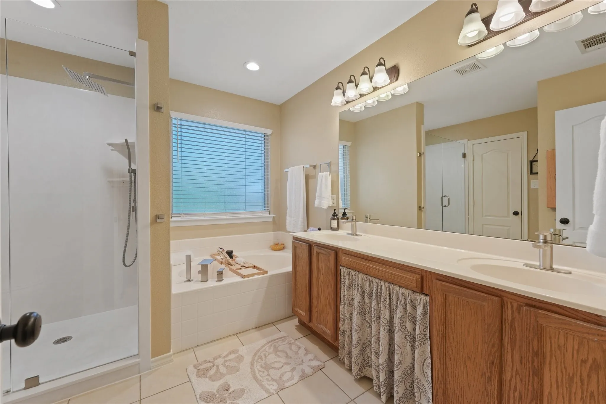 Bathroom featuring a spacious walk-in shower with clear glass enclosure, a built-in soaking tub beneath a window with blinds, and a dual vanity with light-colored countertops and wood cabinetry