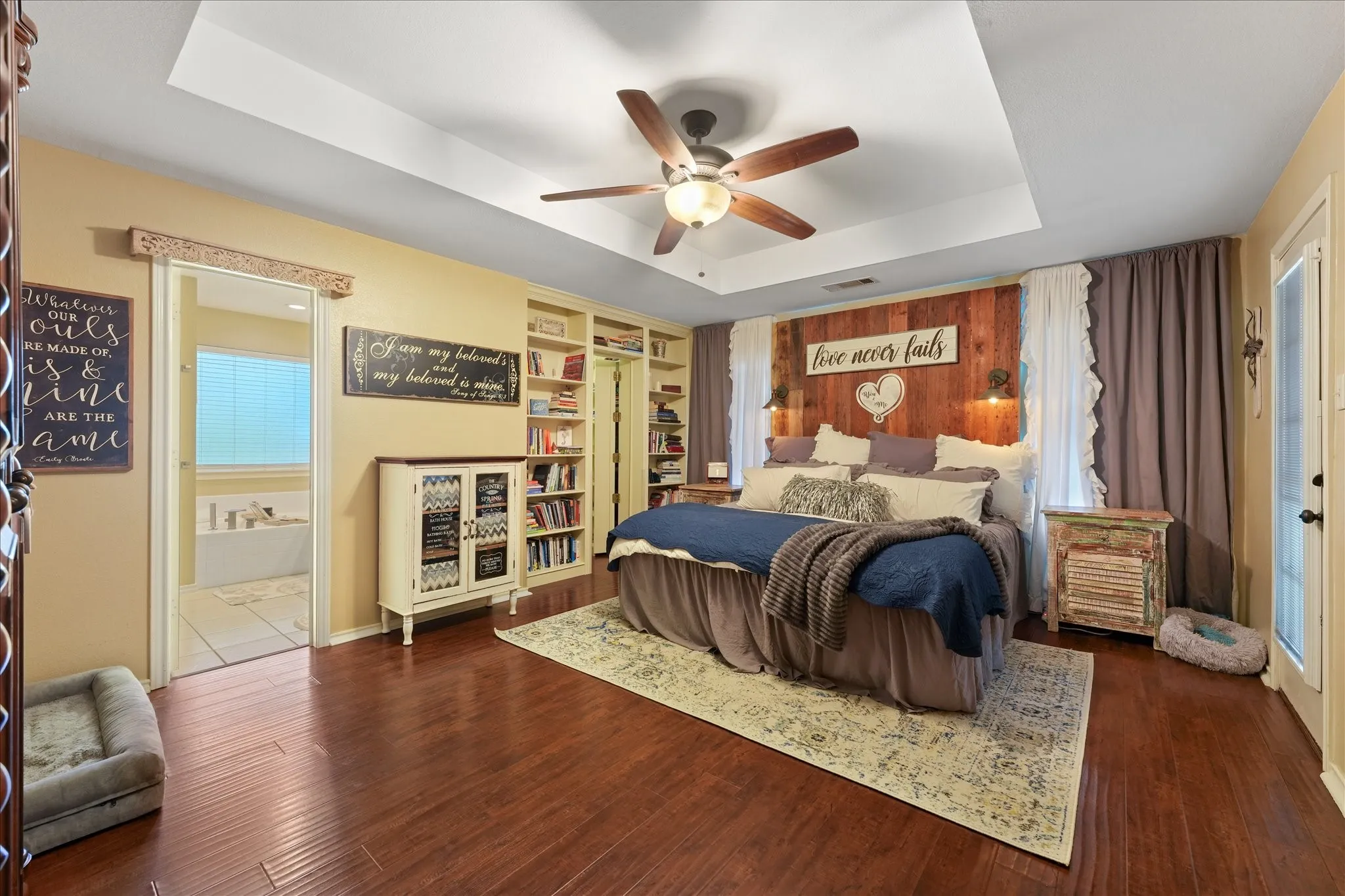 Spacious primary bedroom featuring wood-finish flooring, a tray ceiling with a fan, a wood-paneled accent wall, built-in shelving, hidden walk-in closet and an en-suite bathroom with a soaking tub