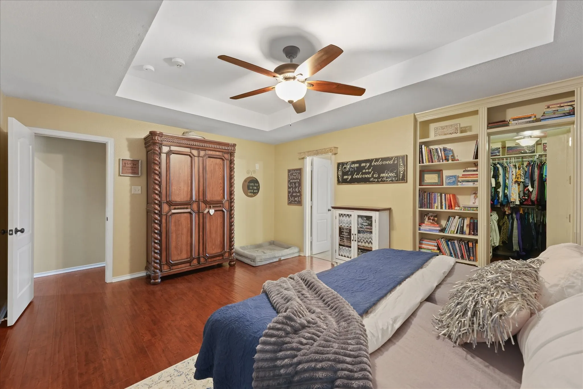 Spacious primary bedroom featuring wood-finish flooring, a tray ceiling with a ceiling fan, a built-in bookshelf, and a hidden walk-in closet with interior lighting