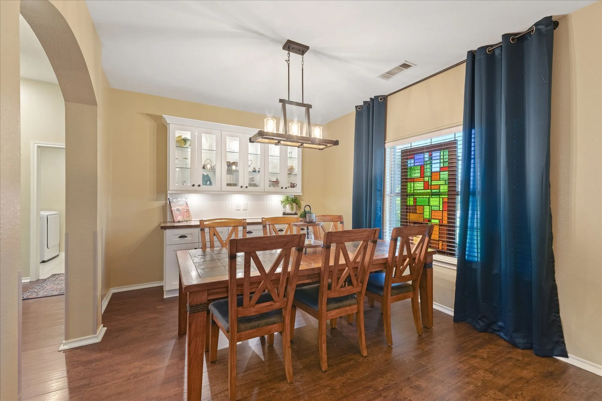 Dining area featuring wood-finish flooring, a built-in display cabinet with glass doors and under-cabinet lighting, a linear chandelier, and a window with a stained glass insert
