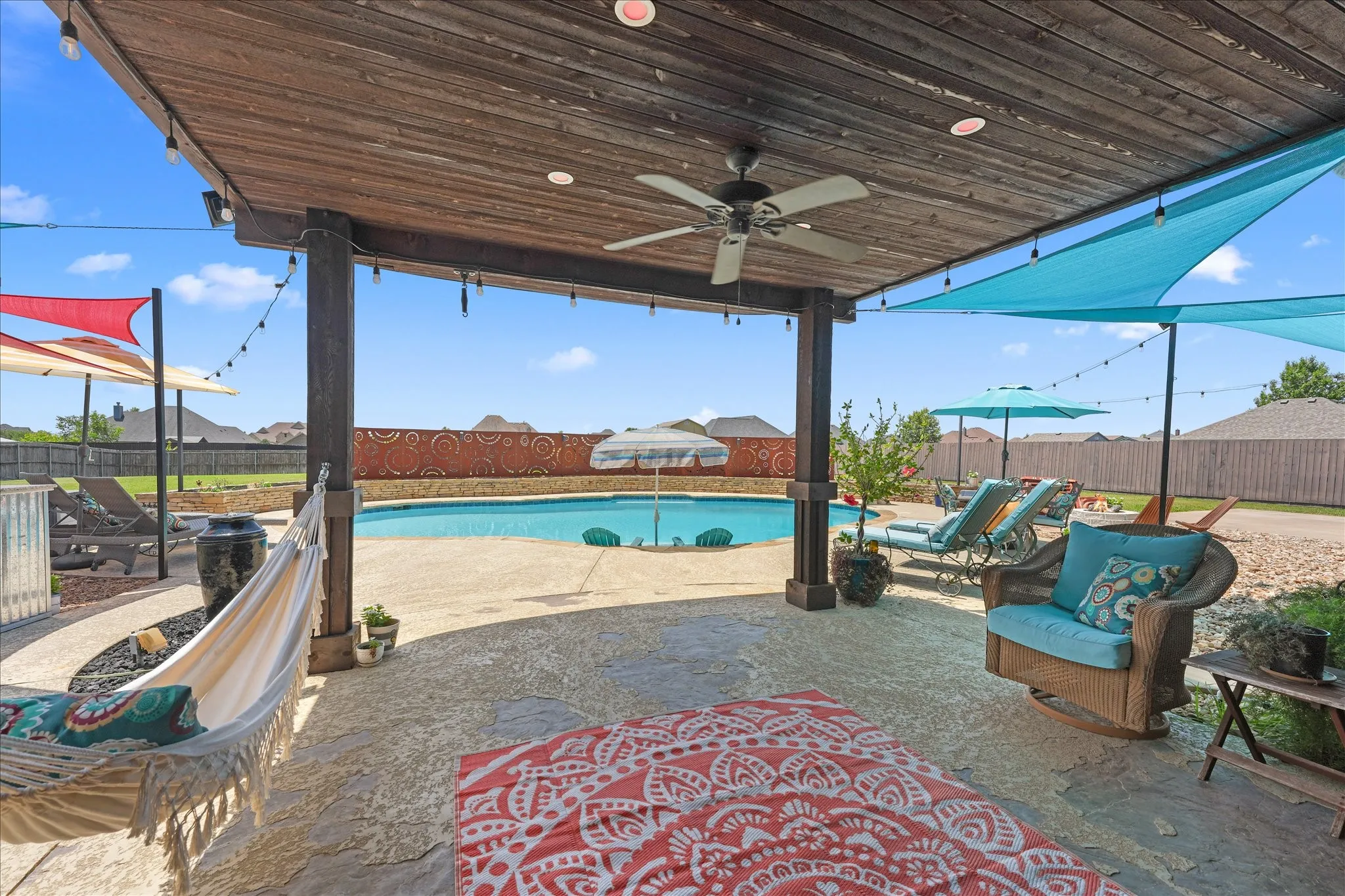Expansive outdoor living area featuring a covered patio with a ceiling fan, string lights, textured concrete surface, an in-ground swimming pool with retaining wall and an art deco steel privacy wall